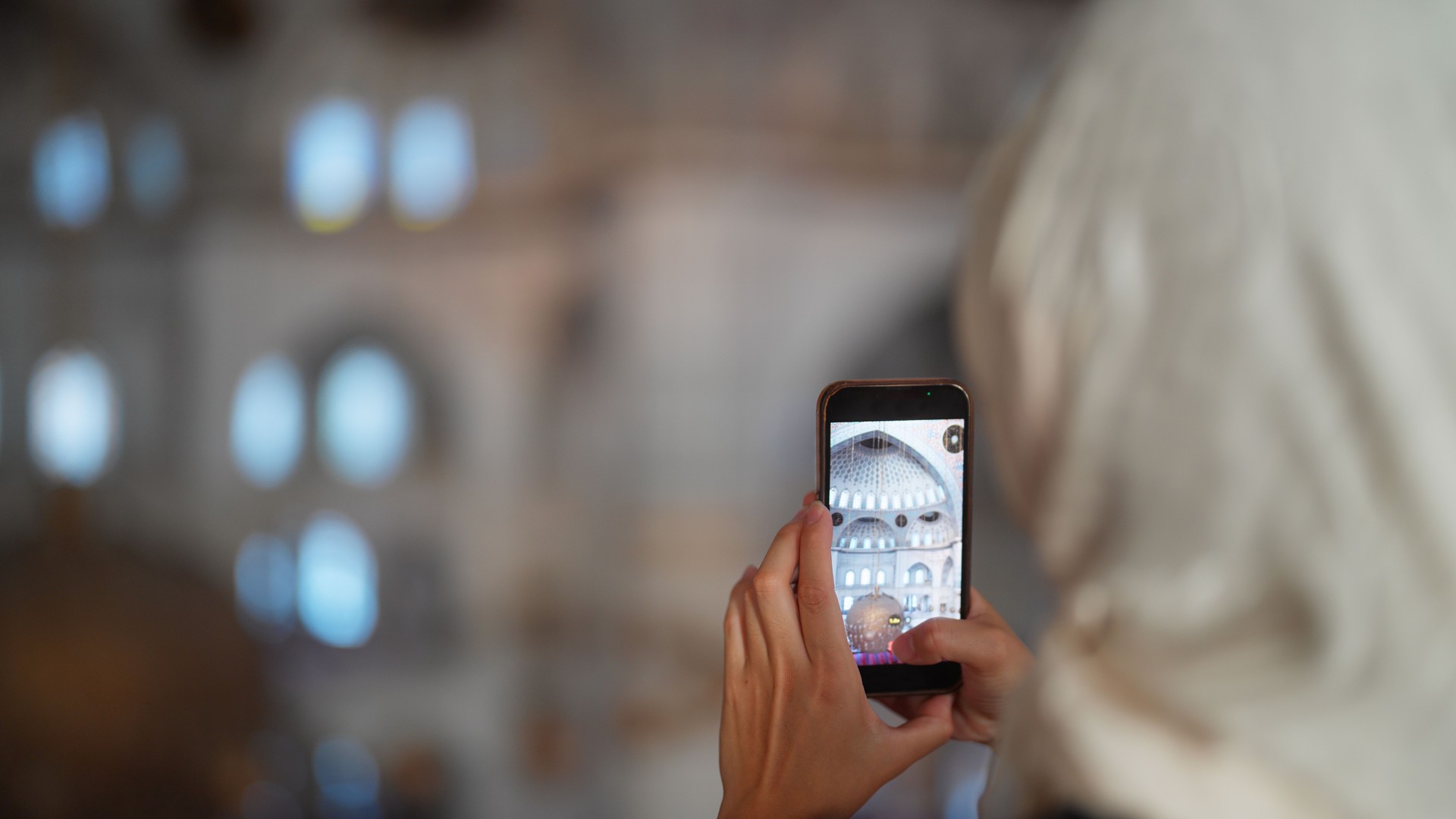 Young female tourist visiting mosque and taking photos and videos with her smart phone